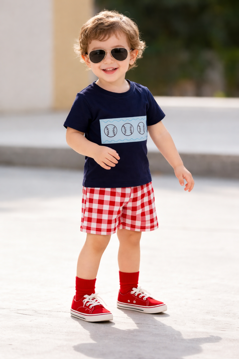 Child wearing a navy blue shirt with a graphic, red checkered shorts, red socks, and red sneakers outdoors.