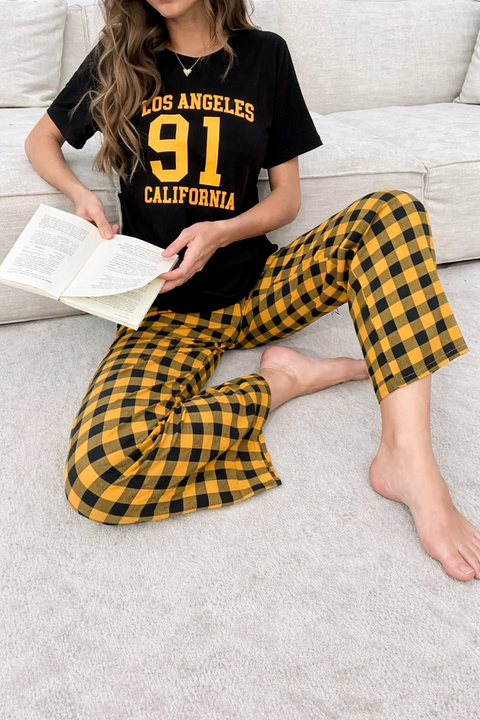 Person wearing a black 'Los Angeles 91 California' t-shirt and yellow plaid pants, sitting on a couch with a book.