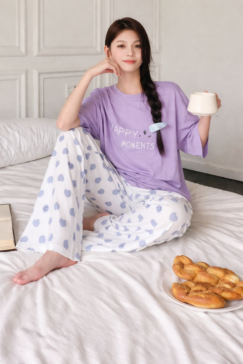 Woman in a purple shirt and patterned pants sitting on a bed with a plate of pastries.