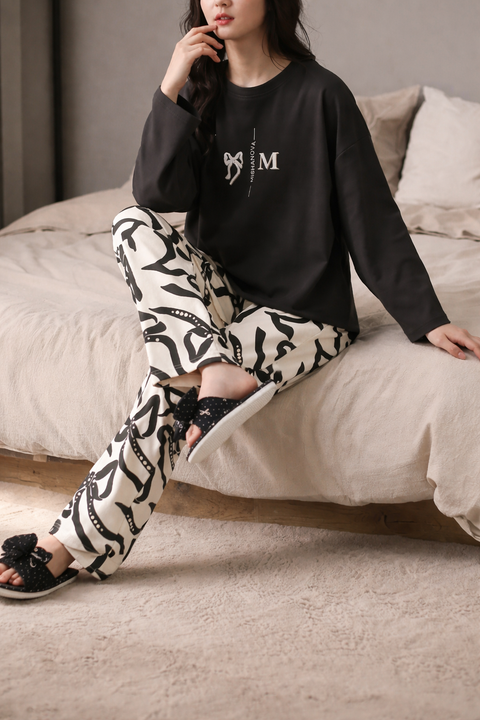 Woman sitting on a bed wearing a black sweatshirt with a logo and patterned pants.